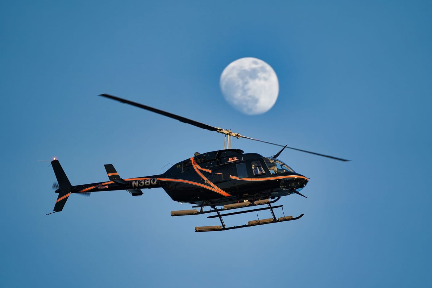 A black and orange private helicopter flying under a blue sky with the moon behind