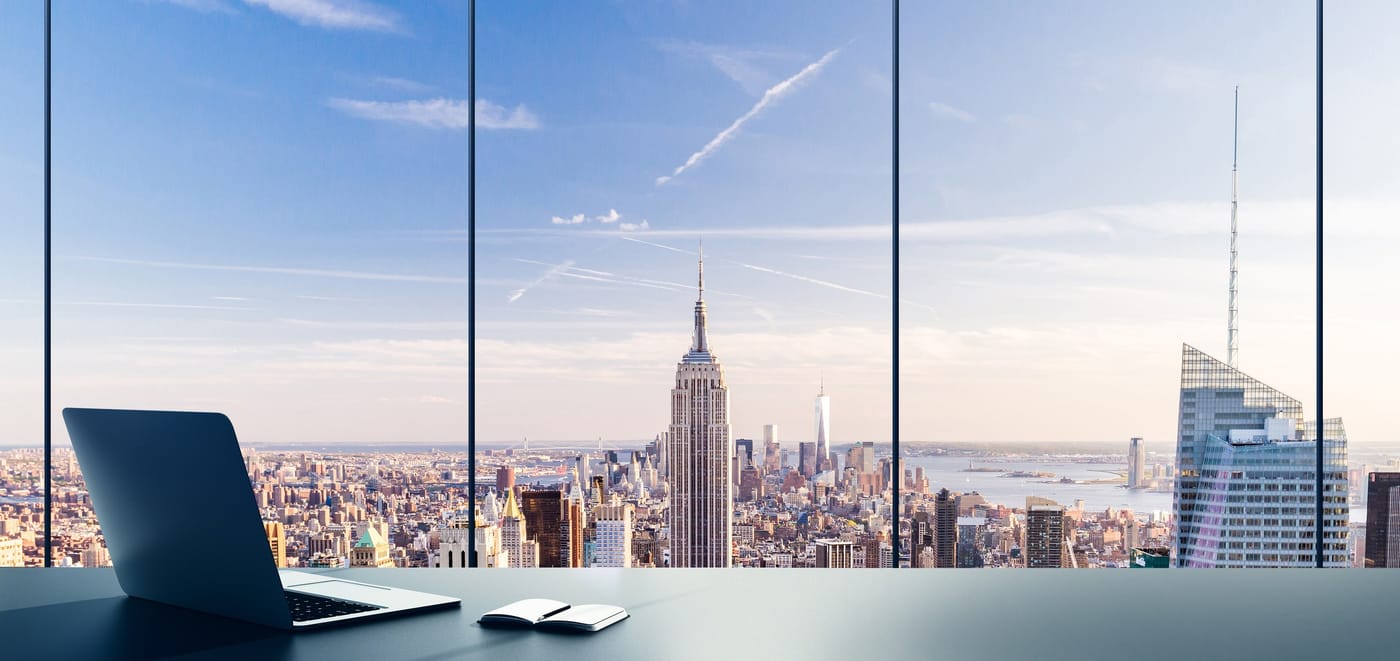 An empty office desk with a laptop, looking out over a city skyline through floor-to-ceiling windows